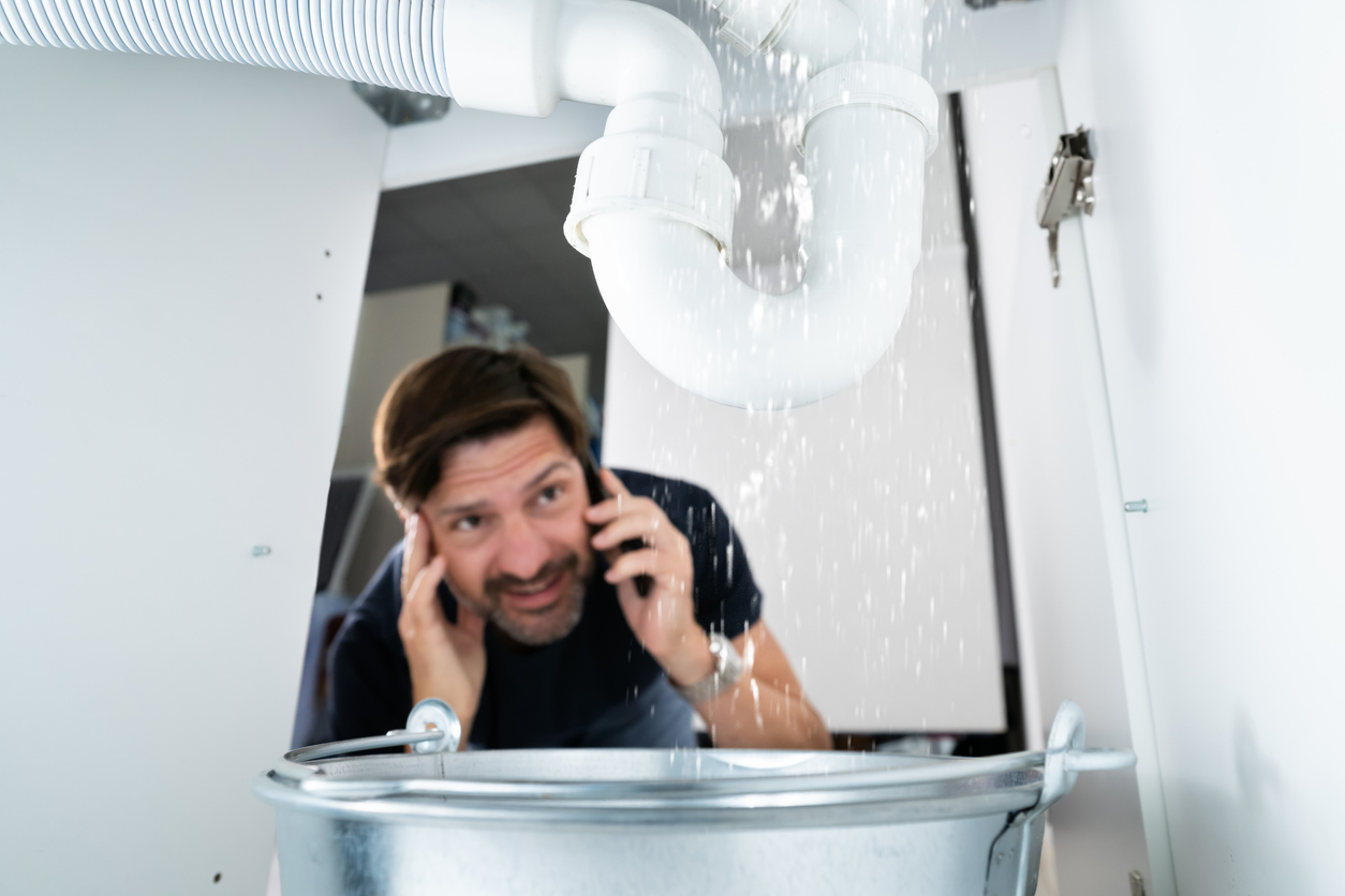 A man crouches under a sink cabinet, holding a phone, as water drips from a pipe into a metal bucket beneath.